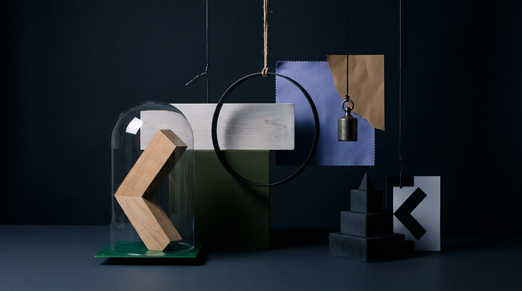 Assorted geometric wooden and metal shapes sit beneath a clear dome beside hanging color swatches in a dark studio.