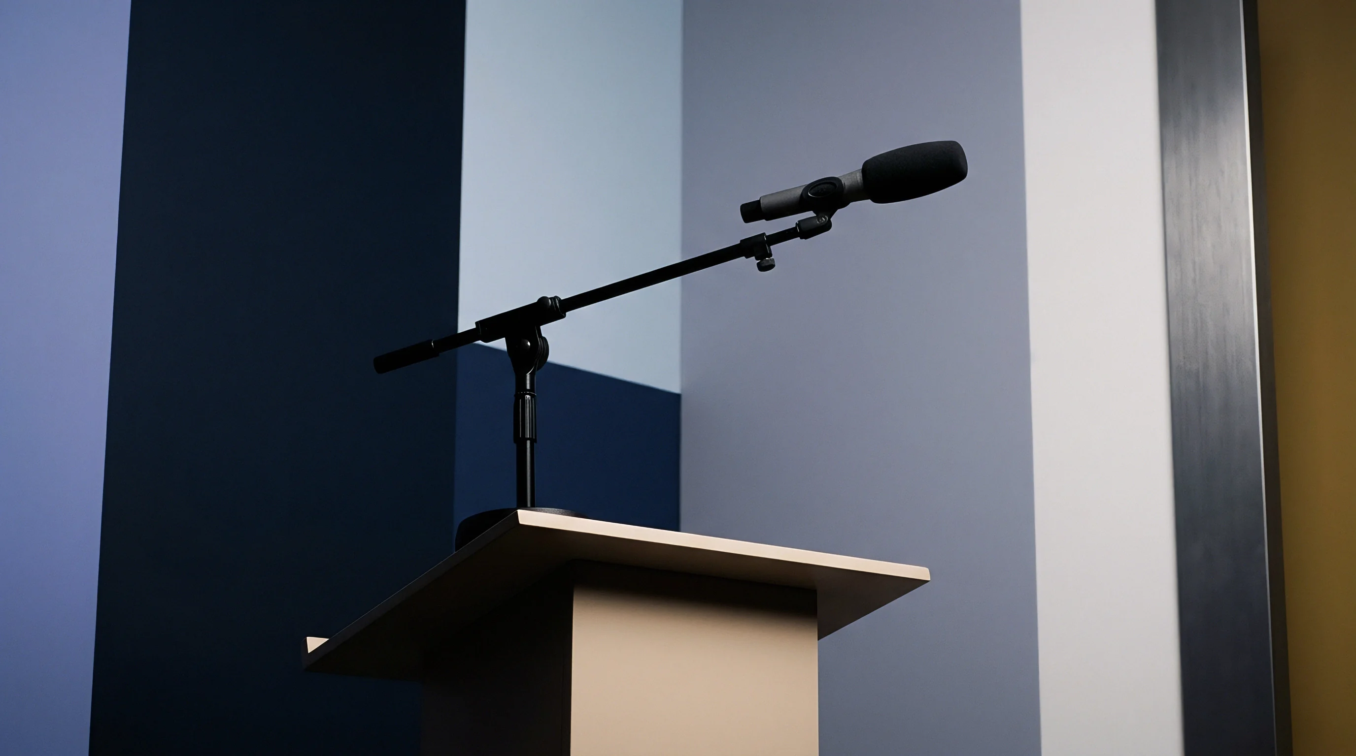 A professional condenser microphone mounted on an adjustable boom arm rests atop a wooden speaker podium positioned before layered geometric architectural panels.