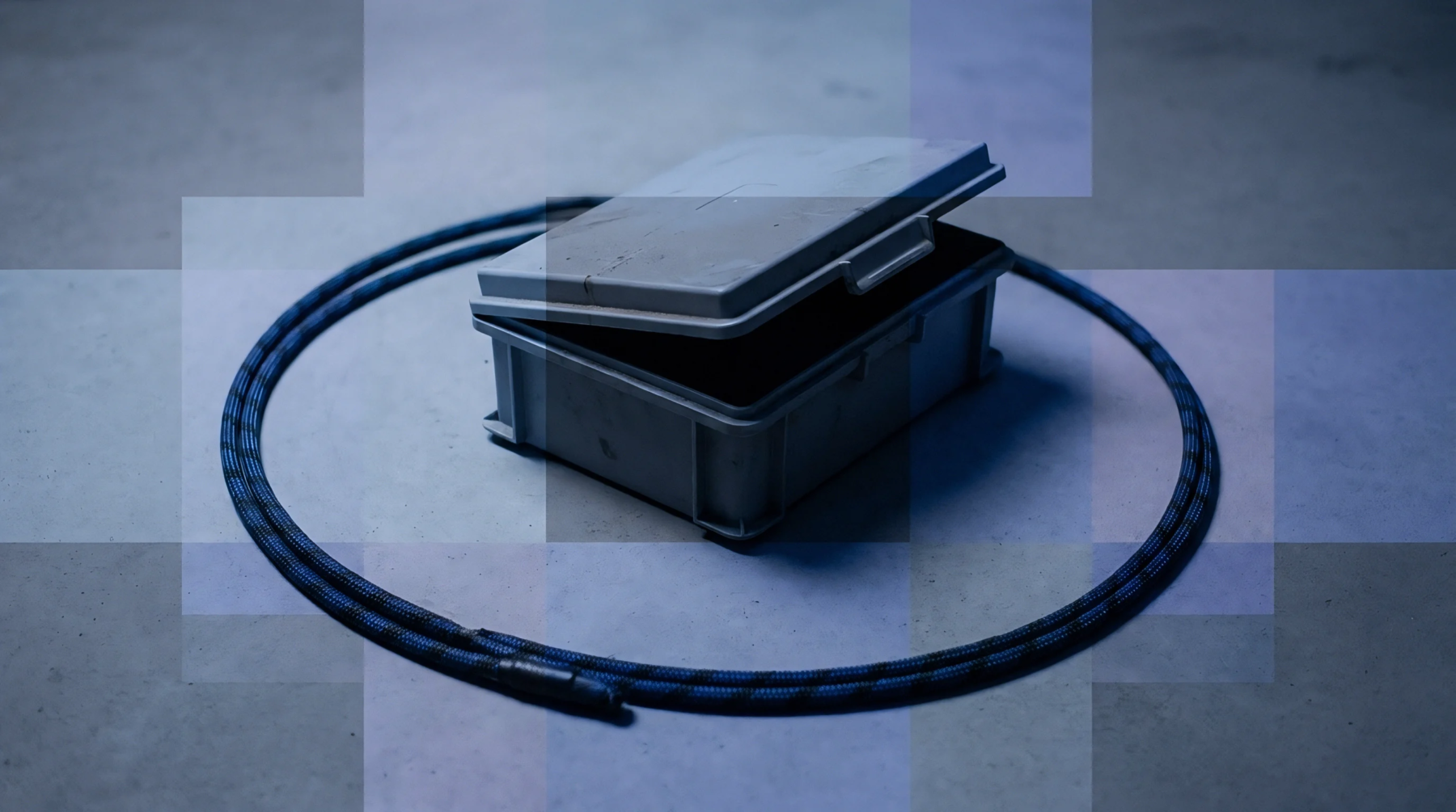 A grey plastic storage container with a partially open lid sits centered on a concrete floor inside a closed loop of braided cable.