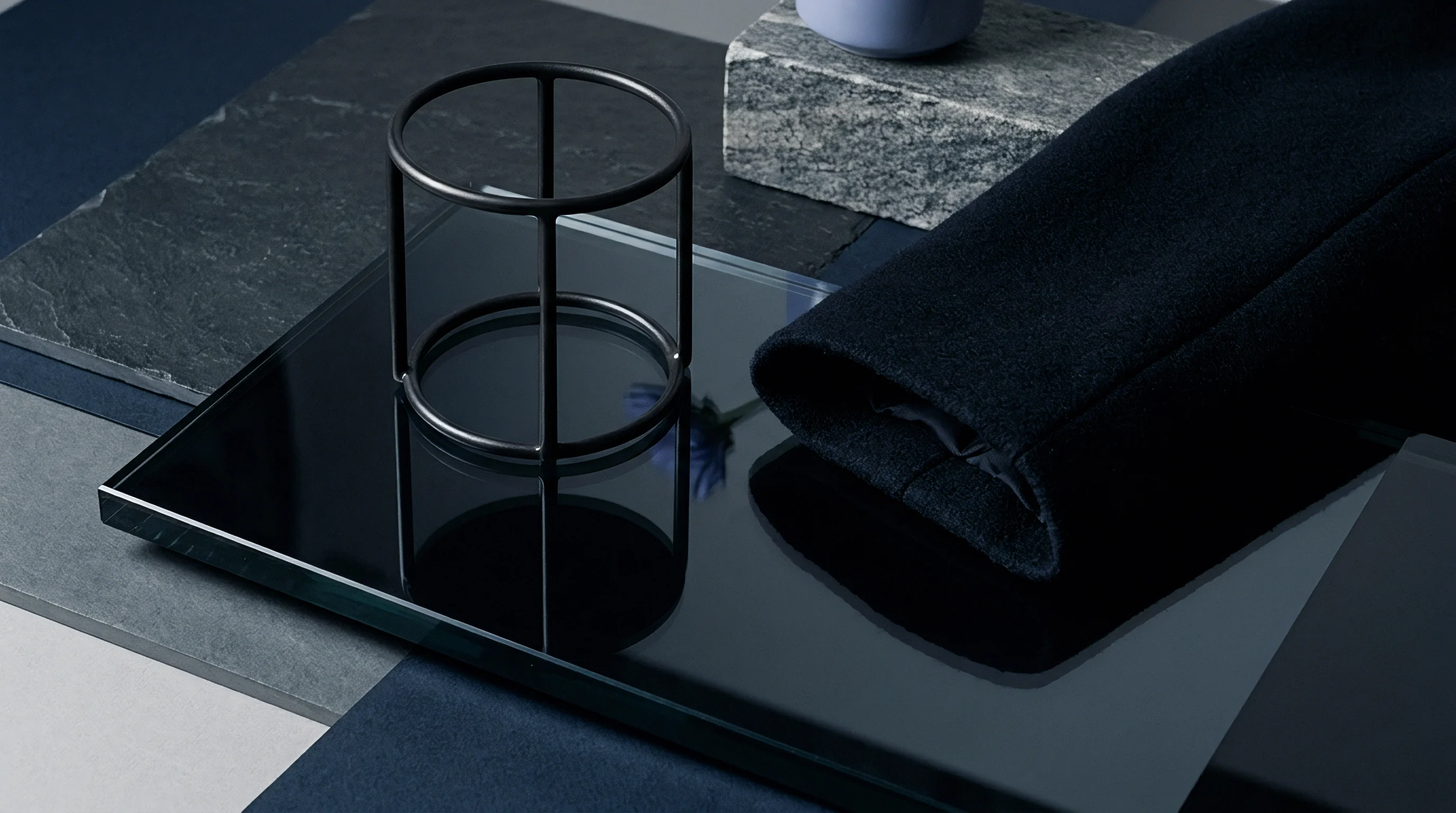 Dark wool textile sleeve rests on a polished glass display tray alongside a minimalist metal wire frame and stone architectural samples.