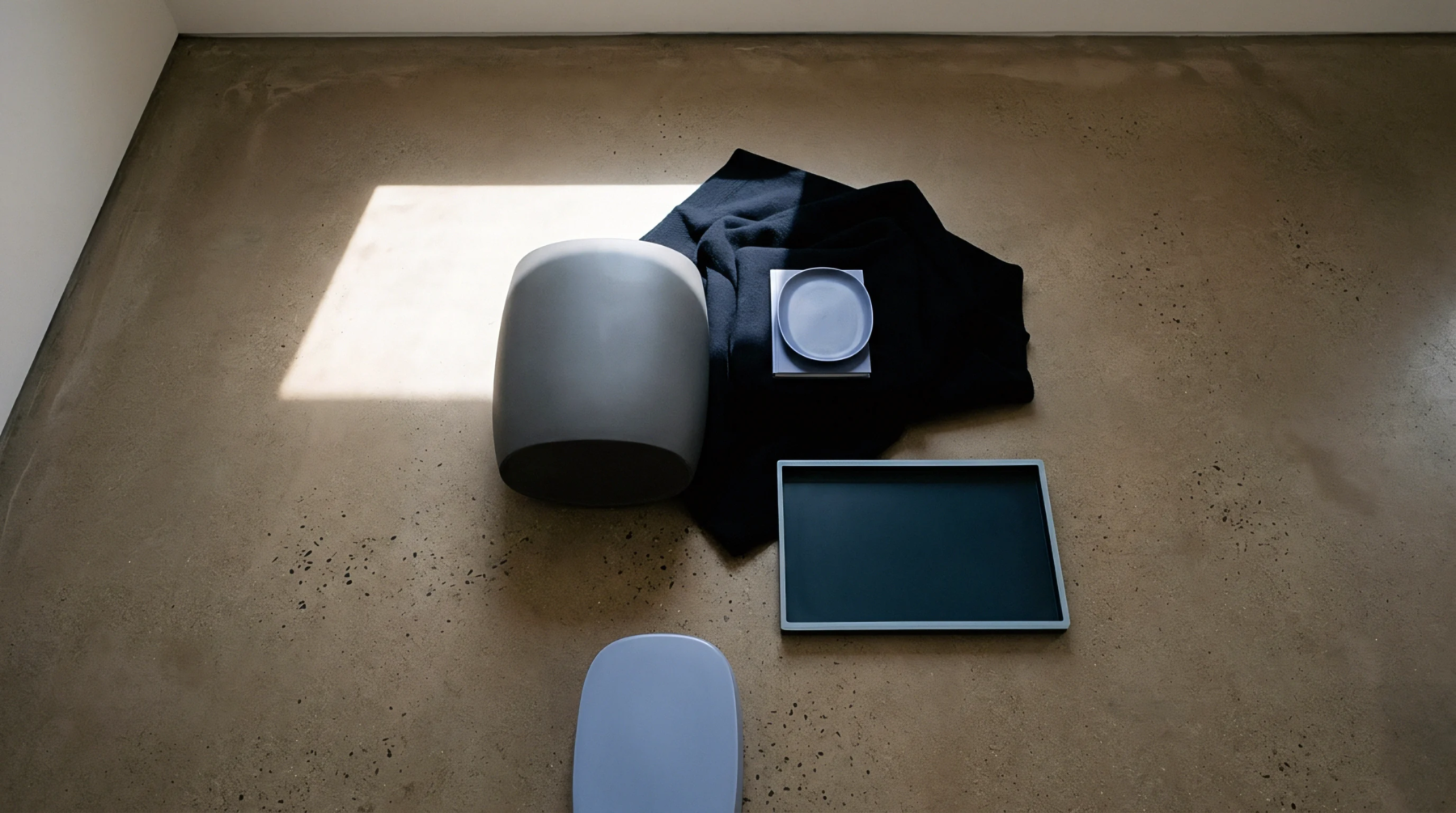 A curated arrangement of industrial materials including a stool, fabric, plate, and tray is presented on a concrete floor under natural light.