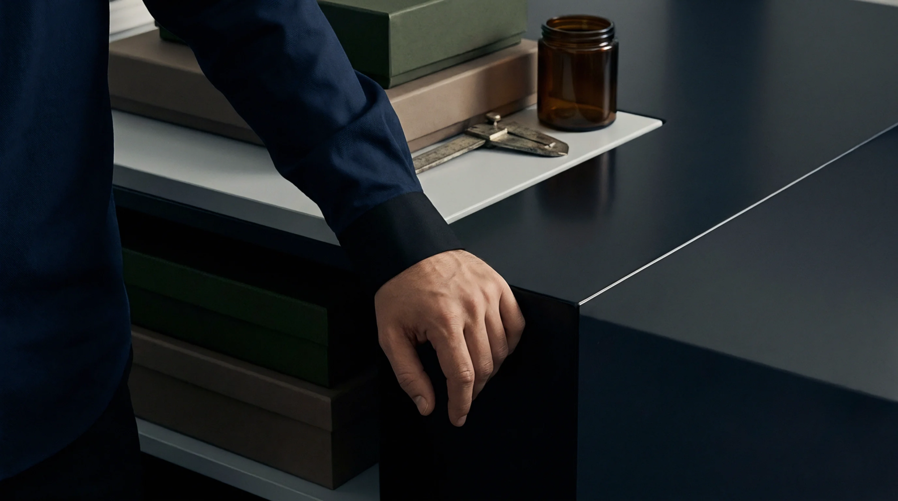 A hand in a navy cuff rests on a dark matte counter beside stacked cardboard boxes and measuring tools.