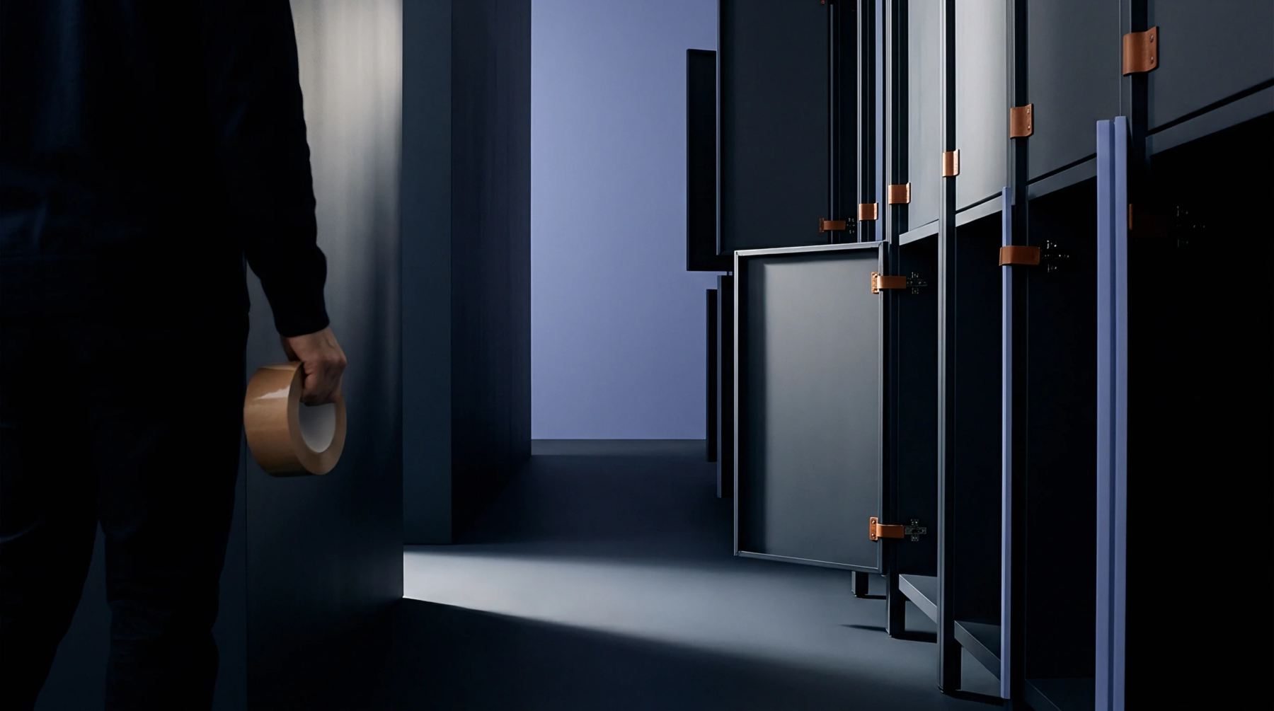 A person in a dark industrial corridor holds a roll of packing tape beside a row of modular storage units.